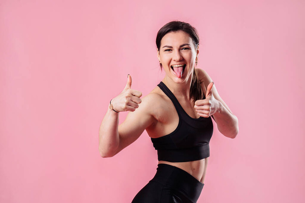 Portret atletycznej, atrakcyjnej muskularnej dziewczyny fitness trener cieszy się życiem i pokazuje szyderstwa i kciuki w studio na różowym tle - Zdjęcie, obraz