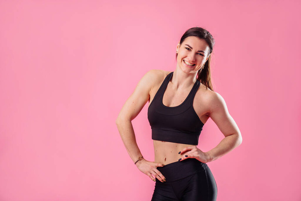 Young pumped up girl coach posing in the studio on a pink background. Dressed in a black top and black pants. Feeling very relaxed and natural, smiles broadly and holds hands on the waist - Фото, изображение