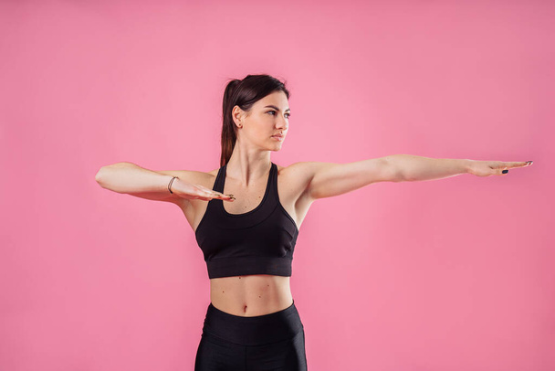 Pumped up sports girl on a pink background in the studio does sports exercises with hands. Smiling and showing her high results by her muscular hands. Fitness for beginners - hands to the side - 写真・画像