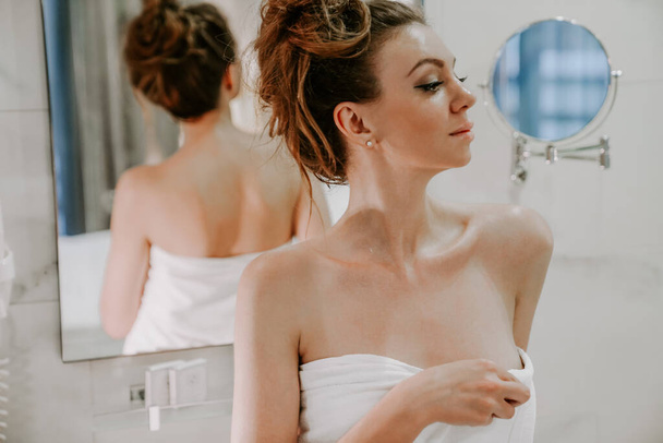 красивая женщина после душа в белом полотенце смотрит в зеркало в ванну. Домашний уход за телом. СПА процедуры
 - Фото, изображение