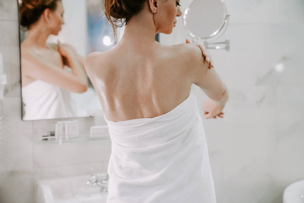 kaunis nainen suihkun jälkeen valkoisessa pyyhkeessä tahraa ihon kermalla. Kotitekoista kehonhoitoa. kylpylähoidot
 - Valokuva, kuva