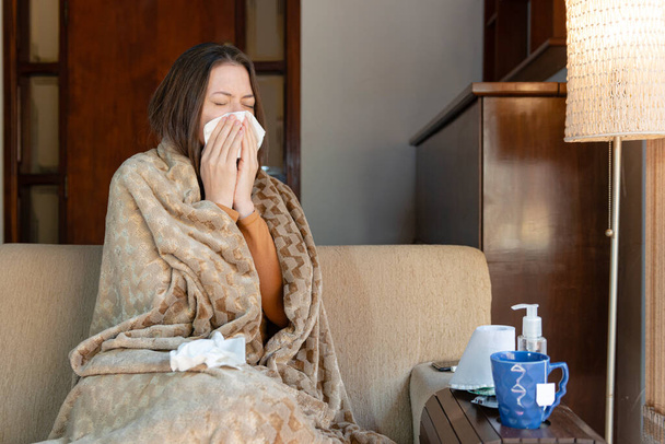 Gyönyörű nő ül a kanapén, kendővel fújja az orrát az intenzív influenza alatt. Feeling beteg koncepció. - Fotó, kép