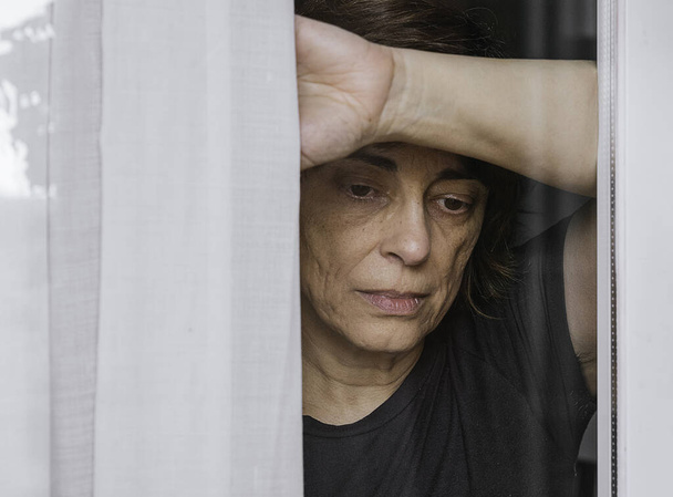 Sad, worried woman looking through the window while there's a quarantine state and she can't go outside - Photo, Image