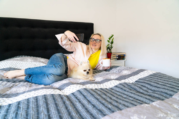 Kobieta korzystająca ze smartfona do rozmowy wideo z przyjacielem lub rodziną. Blogerka z psem siedzącym w sypialni i rozmawiającym z wyznawcami. Webinarium nagrywania Vloggera. Osoba siedzieć na łóżku i patrząc na smartfona - Zdjęcie, obraz