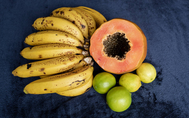 Top kilátás egy friss szeletelt narancs papaya magokkal benne, szeplős banán fürt és három zöld citrom a kék haditengerészet és a fekete háttér. Gyümölcs és étkezési koncepció fotózás - Fotó, kép
