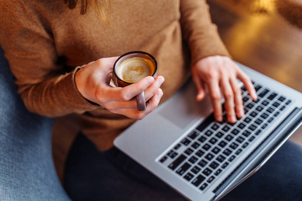 Mądra dziewczyna siedząca i pracująca w biurze z laptopem i filiżanką aromatycznego espresso, cappuccino. Trzymając laptopa na kolanach i ciesząc się przyjemnym aromatem kawy - Zdjęcie, obraz