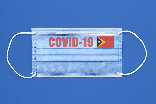 Máscara médica y texto o inscripción Covid-19. Concepto pandémico del Coronavirus. Bandera de Timor Oriental
. - Foto, Imagen
