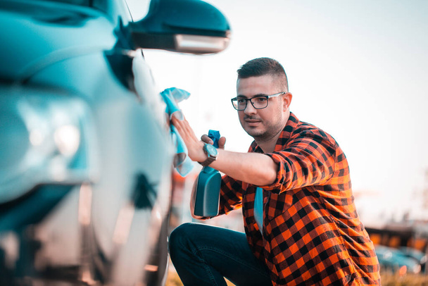 Portrait d'un jeune homme polissant sa voiture avec un chiffon et vaporisant dans une bouteille, à l'extérieur
. - Photo, image
