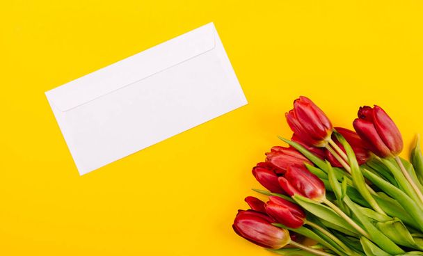 Weißer Geschenkumschlag auf gelbem Hintergrund mit einem Strauß roter Tulpen und einem freien Platz für Text. Musterurkunde oder Einladungskarte. Nahaufnahme. - Foto, Bild