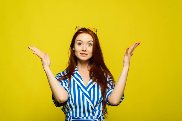 A surprised girl is on the yellow background with glasses on her head. A redhead woman with hands up. - Photo, Image