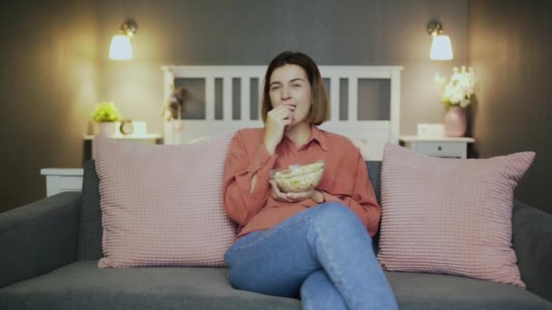 Szczęśliwa młoda kobieta siedząca na kanapie, jedząca popcorn, oglądająca telewizję i śmiejąca się - Materiał filmowy, wideo