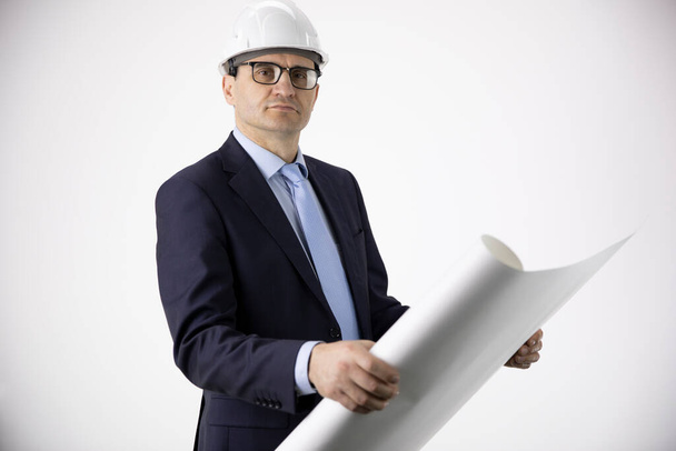 sebevědomý a klidný inženýr v bílé helmě drží kreslení pohledy na fotoaparát - Fotografie, Obrázek