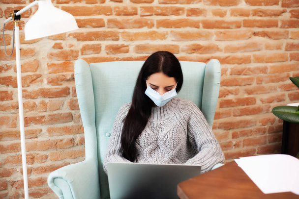 Koruyucu maskeli bir kadın dünya çapında bir virüs salgını sırasında evde dizüstü bilgisayarla çalışıyor. - Fotoğraf, Görsel