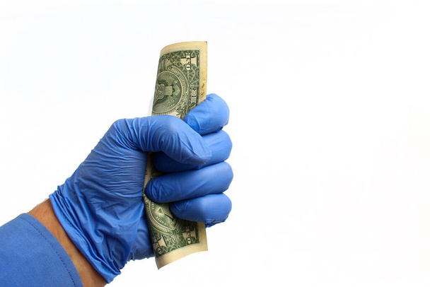 Een hand in een medische handschoen houdt een dollar vast op een witte achtergrond, geïsoleerd. Het concept van de wereldwijde financiële crisis - Foto, afbeelding