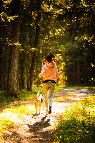 Young woman and dog running together on country in forest. Cheerful female exercising outdoor with her pet. Active people concept. - Foto, imagen