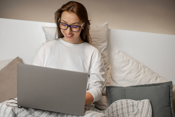 bonita chica de pelo oscuro con gafas se sienta en la cama y sonríe mirando el ordenador portátil
 - Foto, imagen