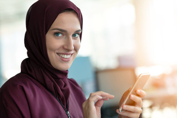 Happy muslim businesswoman in hijab at office workplace. Smiling Arabic woman working on laptop and talking on smartphone - Foto, immagini