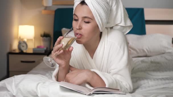 Girl drinking champagne and reading book. Attractive woman in bedroom in white bathrobe with towel on her head lies in bed drinks wine and reads book, turns over pages. Close-up - Imágenes, Vídeo