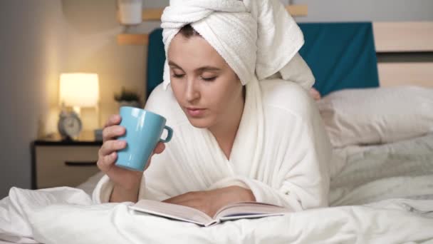 Girl drinking coffee or tea and reading book. Attractive woman in bedroom in white bathrobe with towel on her head lies on stomach drinks hot coffee or tea and reads book, turns over pages. Close-up - Video