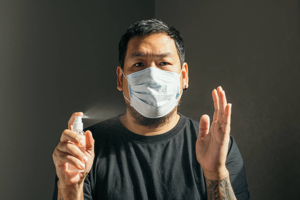 Ιός του κερατοειδούς. Άντρας φορώντας ιατρική προστατευτική μάσκα καθαρισμού των χεριών με ψεκασμό αποστειρωτήρα στο σπίτι για την πρόληψη του ιού του Coronavirus, Covid-19, γρίπη. Μπουκάλι. Ιός και προστασία της ασθένειας. - Φωτογραφία, εικόνα