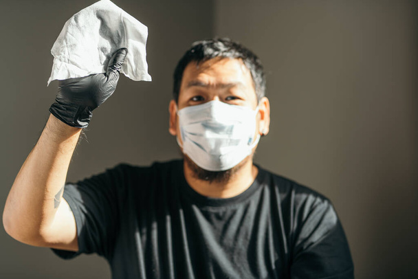 Coronavirus. Mann mit medizinischer Schutzmaske und Latexhandschuh, der Papiertücher im Haus zeigt, um Coronavirus, Covid-19, Grippe vorzubeugen. Sprühflasche. Viren- und Krankheitsschutz. - Foto, Bild