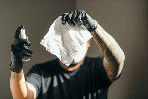 Coronavirus. Mann mit medizinischer Schutzmaske und Latex-Handschuh mit Papiertücher und Sprühflasche im Haus, um Coronavirus, Covid-19, Grippe vorzubeugen. Sprühflasche. Viren- und Krankheitsschutz. Fucus am Scheideweg. - Foto, Bild