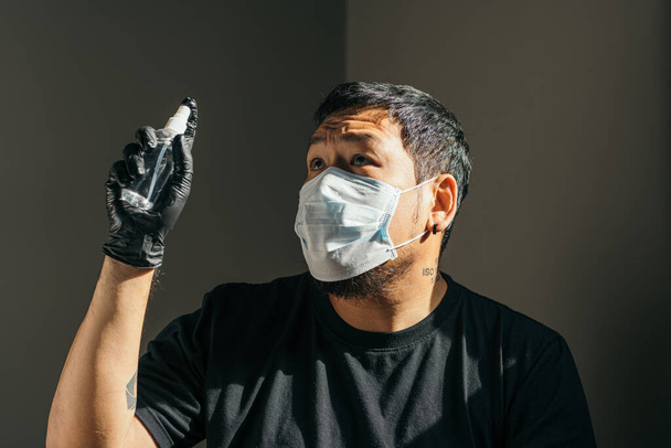 Coronavirus. Man wearing in medical protective mask and black rubber glove spraying with sanitizer spray in house to prevent Coronavirus, Covid-19, flu. Spray bottle. Virus and illness protection. - Photo, Image