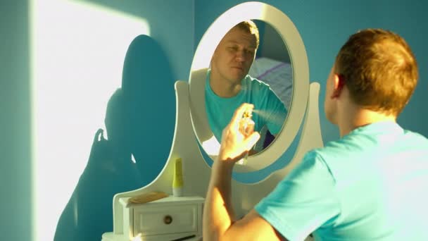 Joven hombre atractivo se sienta delante de un espejo blanco y rocía perfume sobre sí mismo
 - Imágenes, Vídeo