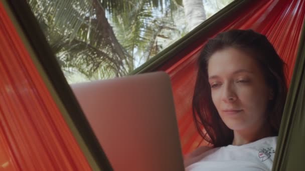 Természetes nő dolgozik notebook szabadban ül függőágyban zöld pálmafák háttér. Laptopot használó lány a friss levegőn, lassítva zárja le a felvételt. Szabadúszó távmunka nyári vakáció életmód - Felvétel, videó