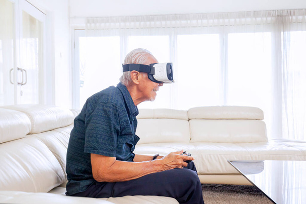 Side portrait of old unidentified man wearing vr glasses, while playing it with a controller happily in his living room - Foto, afbeelding