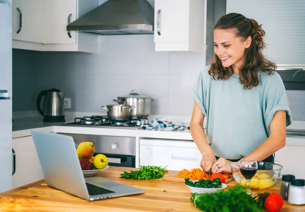 Happy beautiful young Woman enjoying cooking looking for a recipe on the laptop in the kitchen Laptop on kitchen table and cooking girl. Food blogger concept - 写真・画像