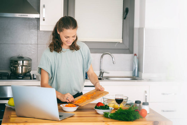 Happy beautiful young Woman enjoying cooking looking for a recipe on the laptop in the kitchen Laptop on kitchen table and cooking girl. Food blogger concept - Foto, immagini