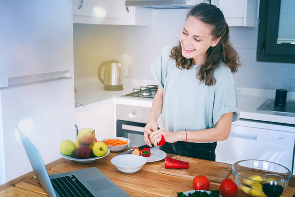 Happy beautiful young Woman enjoying cooking looking for a recipe on the laptop in the kitchen Laptop on kitchen table and cooking girl. Food blogger concept - Fotografie, Obrázek