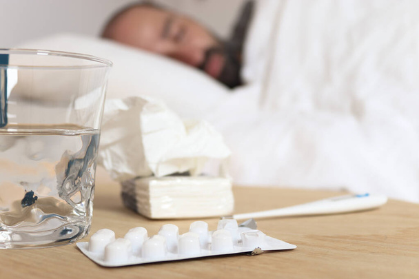 Homem doente na cama com lençóis brancos. Acima da mesa estão comprimidos, lenços, um termómetro e um copo de água.
 - Foto, Imagem
