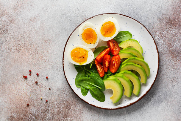 Gesundes vegetarisches Salatfrühstück. Spinatblätter, Tomate, Avocado und gekochtes Ei auf grauem Teller und grauem Betonboden. Buddha Schale vorhanden. Ansicht von oben. - Foto, Bild
