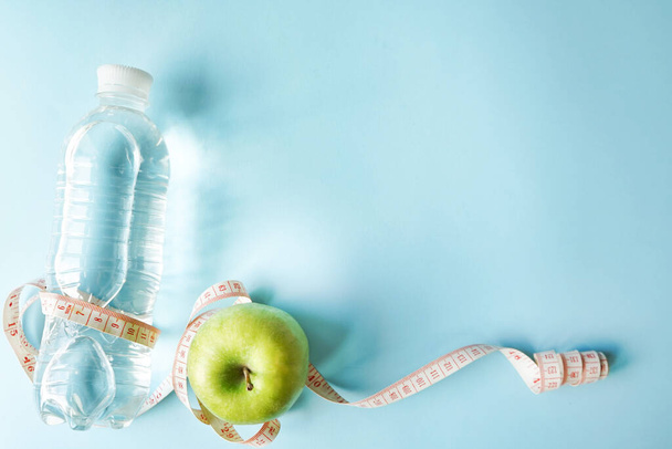 Diet symbol flat lay one meter ribbon and green apple and a bottle of water. blue background with copy space. health sport concept - 写真・画像
