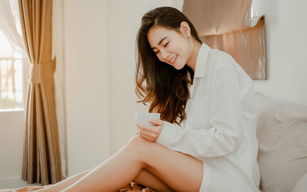 Молодая женщина, живущая дома, отдыхает и пьет чашку горячего кофе в спальне на отдыхе. Азиатский, азиатский, расслабляющий, одинокий, технология, концепция образа жизни. - Фото, изображение