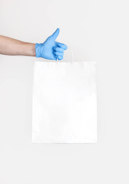 Concept van levering van goederen tijdens quarantaine. Jongeman in blauwe handschoenen houdt witte papieren zak in zijn handen op een witte achtergrond. Levering tegen Coronavirus 2019-nCov in pandemie Contactloze levering. - Foto, afbeelding