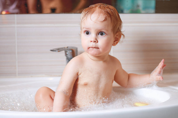 Ευτυχισμένο το γέλιο βρέφος νήπιο λαμβάνοντας ένα μπάνιο παίζοντας με φυσαλίδες αφρού. Μπάνιο και πλύσιμο του μικρού παιδιού. Η έννοια της φροντίδας και της υγιεινής των παιδιών. - Φωτογραφία, εικόνα