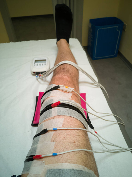 Menschliches Bein mit Muskelelektrostimulationsgerät und Eisbeutel während Rehabilitationsübungen auf dem Bett nach einer Knieoperation. - Foto, Bild