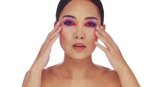 Счастливая полуобнаженная азиатка с ярким макияжем глаз моды касается ее лица, глядя на камеру изолированы на белом фоне в студии
 - Кадры, видео
