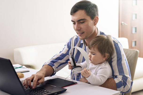 膝の上の赤ん坊と縞模様のシャツの黒い髪の男隔離されたリモートワークヘッドフォンで音楽を聴く家庭での安全な仕事幸せな父親の概念 - 写真・画像