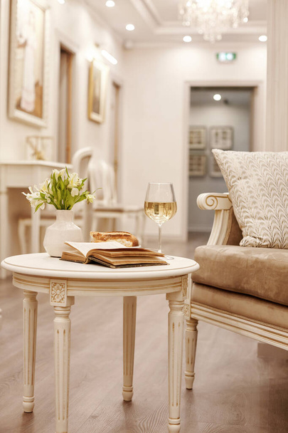 Egy nyitott évjárat könyv, egy pohár fehér bor és ropogós sütemények ülnek egy vintage asztalon a világos, hangulatos nappaliban - Fotó, kép