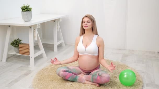 A young beautiful pregnant Caucasian woman sits on the soft carpet of the house. He is engaged in fitness and doing exercises. Sportswear. - Imágenes, Vídeo