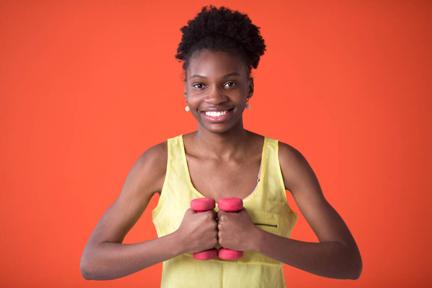 beautiful young african woman on a red background in a yellow t-shirt with dumbbells - Photo, image