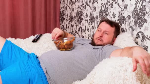 tlustý muž s velkým břichem leží na gauči s rychlým občerstvením a je znuděný před televizí s dálkovým ovládáním v ruce, zblízka. - Záběry, video