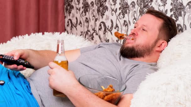 Fat man met baard met een dikke buik ligt op de bank met fastfood, drinkt bier en mist de TV met de afstandsbediening in zijn hand, close-up. - Video