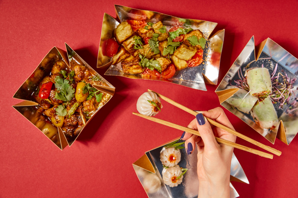 Вид сверху женщины, держащей в руках палочки с тушеной булочкой рядом с вкусной китайской едой в коробках на красном фоне - Фото, изображение