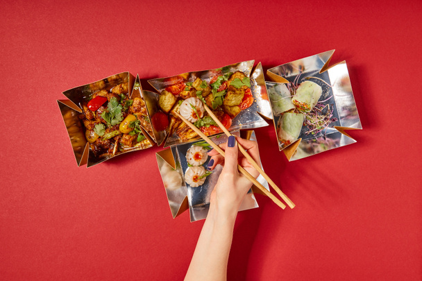 top view of woman holding chopsticks with steamed bun near prepared chinese food in takeaway boxes on red - Photo, Image