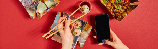 Панорамный снимок женщины, держащей в руках палочки-выручалочки с тушеной булочкой и смартфон с экраном-бланком рядом с вкусной китайской едой в коробках на красном фоне - Фото, изображение